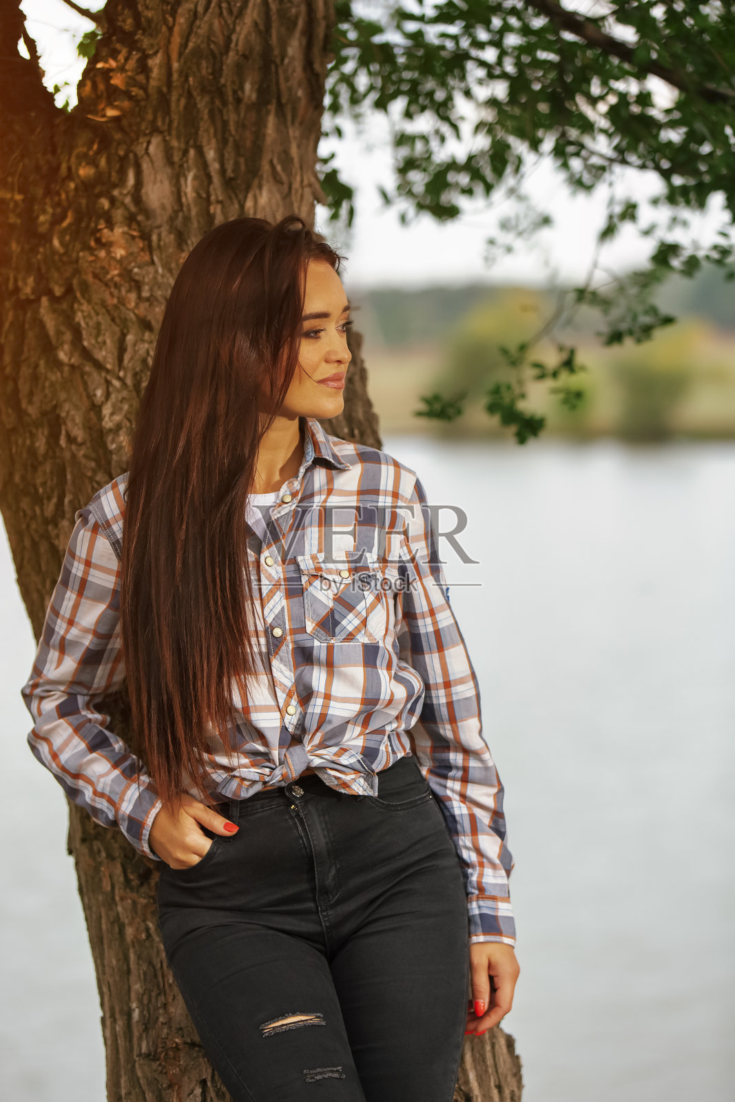 一个穿着格子衬衫和牛仔裤的女人倚在河边的一棵树上照片摄影图片