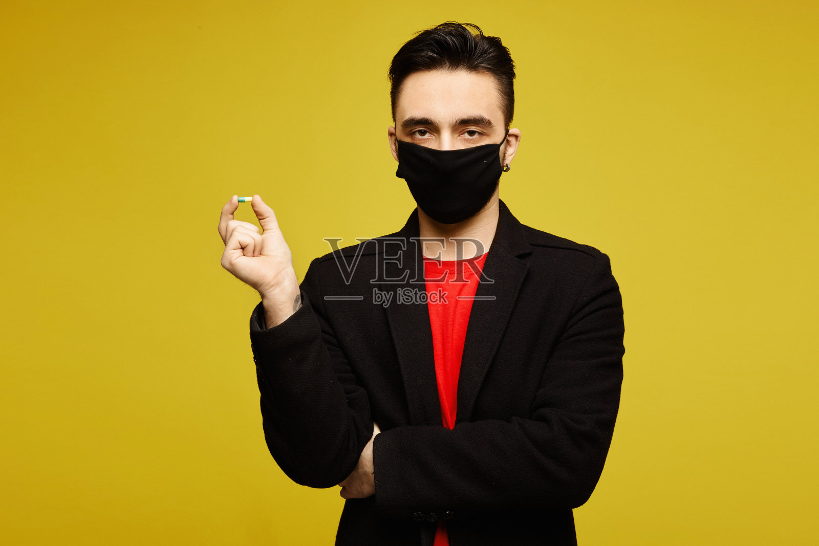 身着黑色外套,戴着黑色防护面具的帅气男子手里拿着一颗药丸,在黄色背景下摆姿势,与世隔绝。医学病毒。医疗保健的生活方式。照片摄影图片