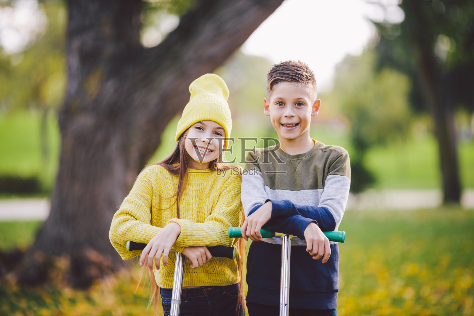 两个孩子,白人男孩和女孩双胞胎摆姿势与滑板车在秋季公园。快乐的孩子们在公园里骑滑板车。孩子的运动。户外活动弟弟妹妹青少年。快乐的童年照片摄影图片