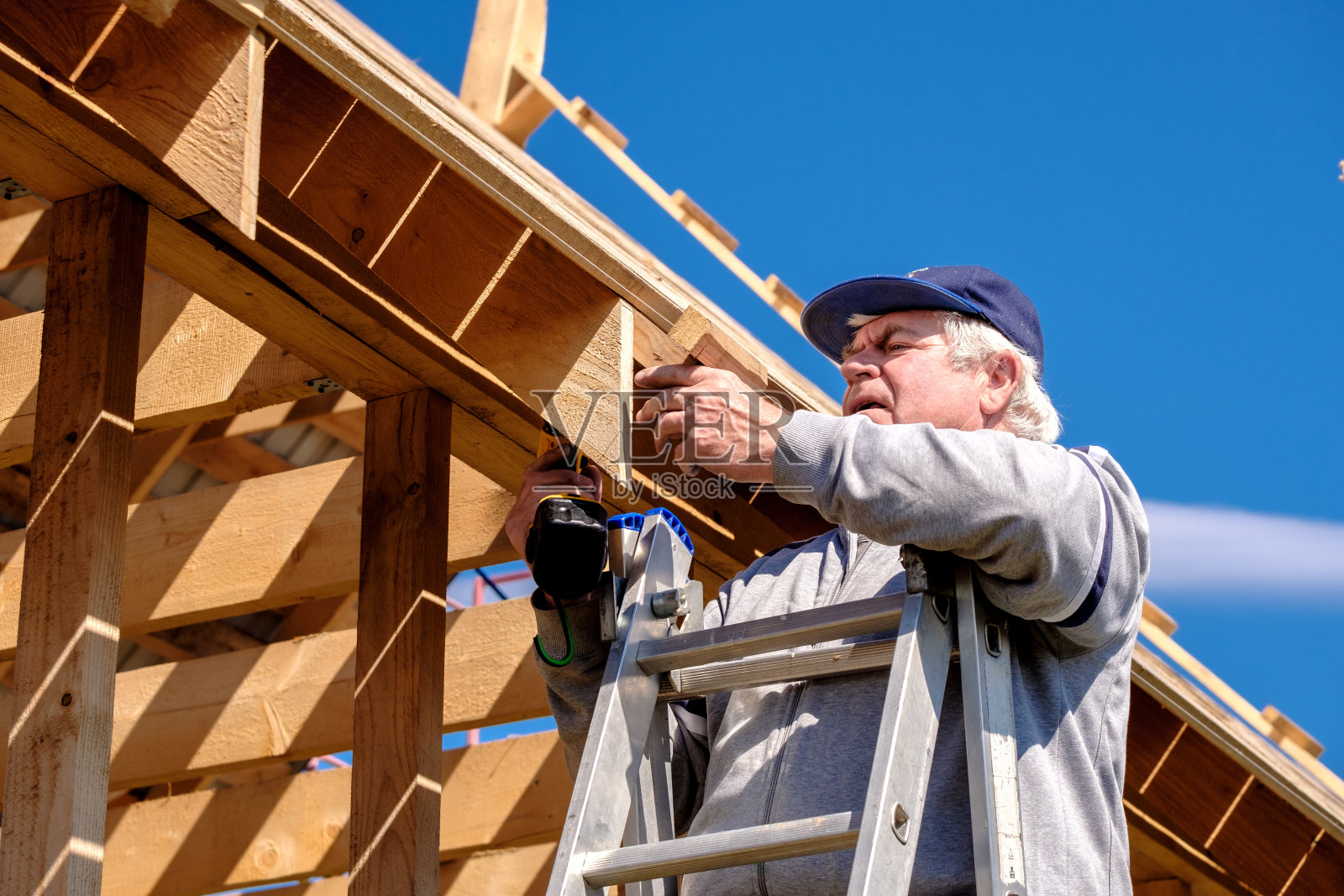 一位白发苍苍的资深建筑工人在一个春日的蓝天下,用一个无绳螺丝刀收集了一座乡村木屋的框架。照片摄影图片
