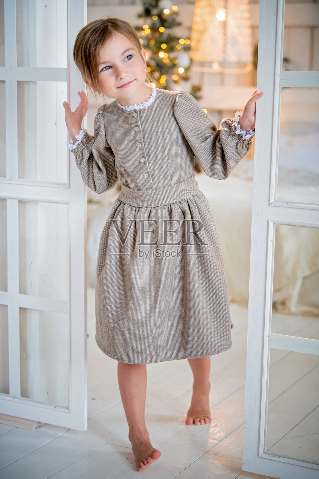 在一个明亮的室内女孩高兴地看着门外,笑了。透过玻璃可以看到一棵圣诞树。照片摄影图片