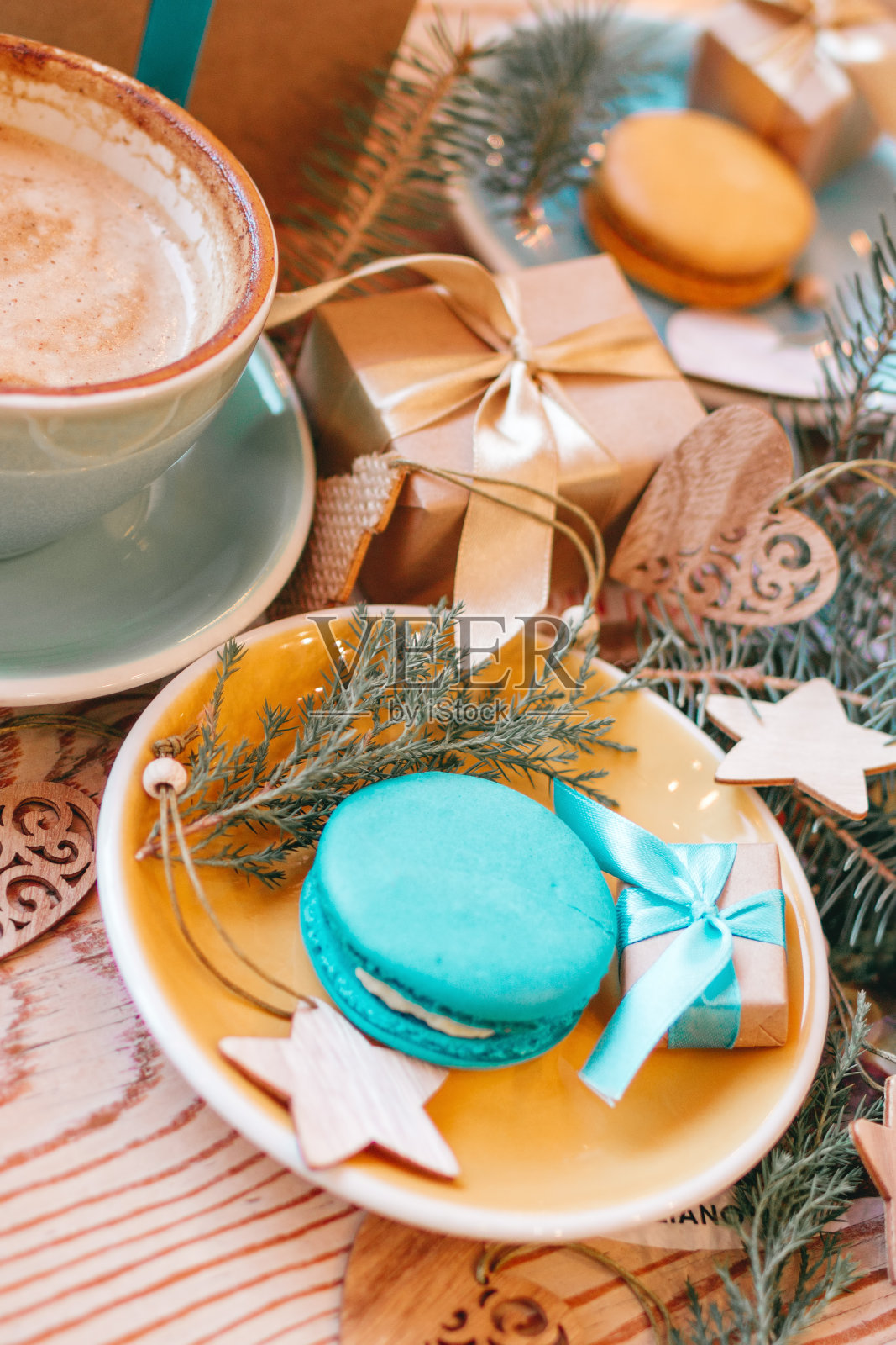蓝色杯子配卡布奇诺,蓝色和黄色的托盘上放着甜马卡龙,木制背景上有圣诞礼物,圣诞装饰品和针叶树枝,俯视图。照片摄影图片