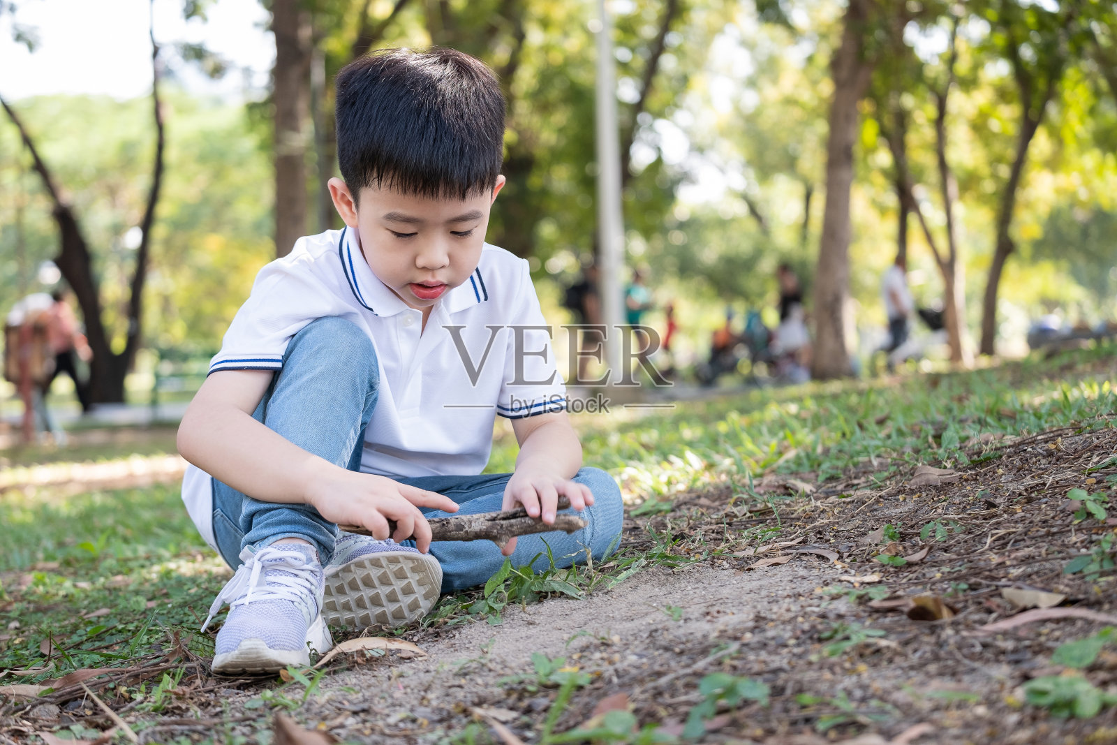 一个亚洲男孩坐在公园里,用树枝或树枝在地上画画。穿着白色t恤和牛仔裤的孩子。户外和家庭活动。创意和肖像概念照片摄影图片