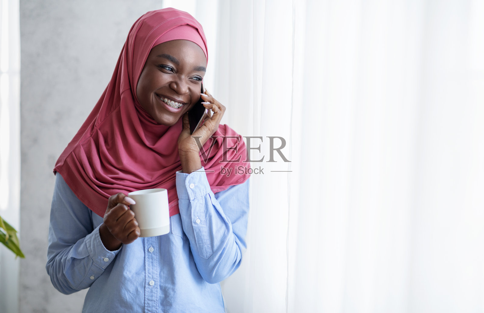 微笑的黑人穆斯林女士在窗边用手机和咖啡放松照片摄影图片
