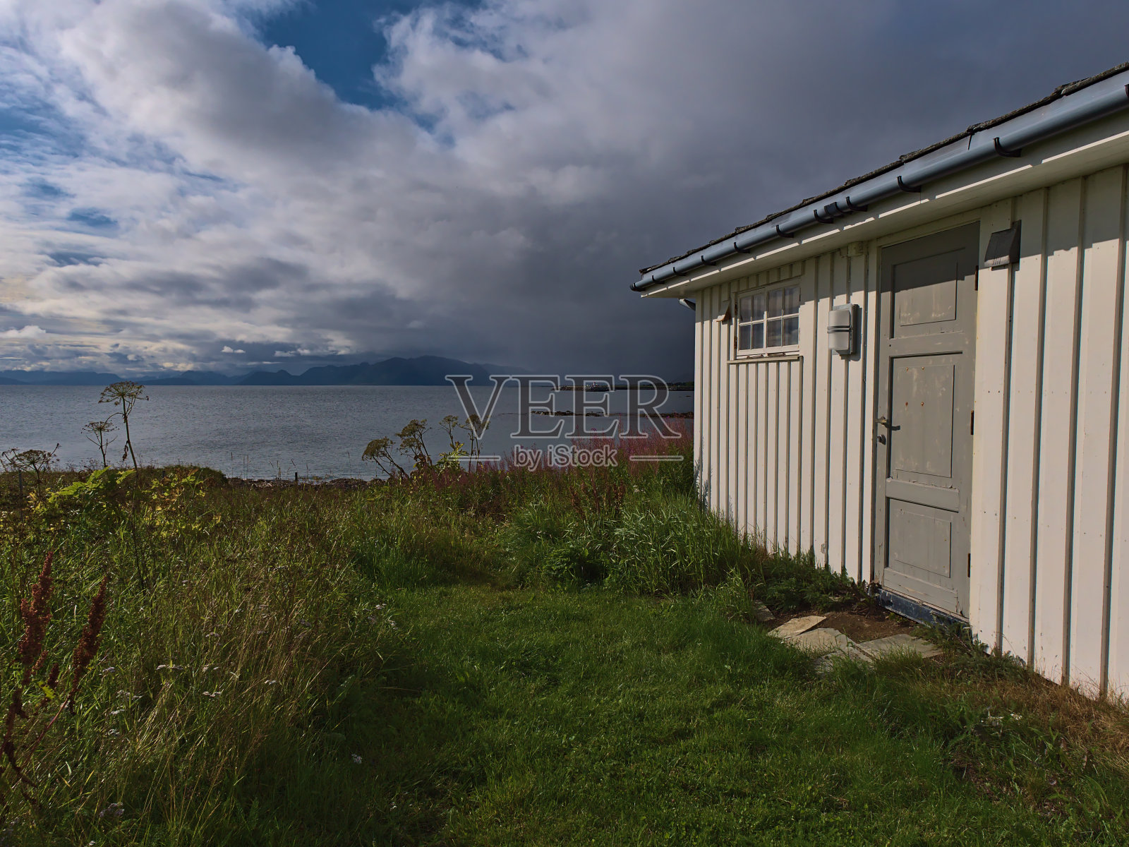 白色粉刷的木屋,有门和高草草地,在Andøya岛Andfjord海岸附近的村庄Dverberg, Vesterålen,挪威,在阳光明媚的一天崎岖的山脉。照片摄影图片