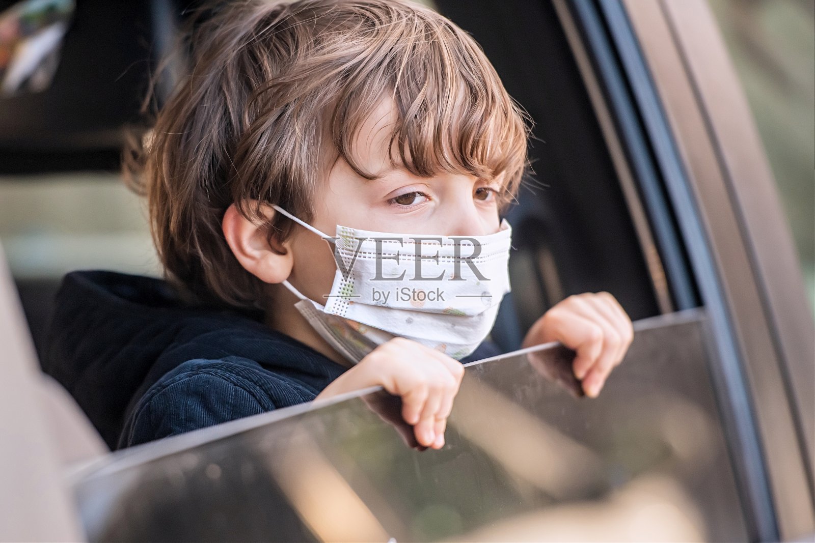一个戴着防护面罩的小男孩在上学的路上,透过车窗看向别处照片摄影图片