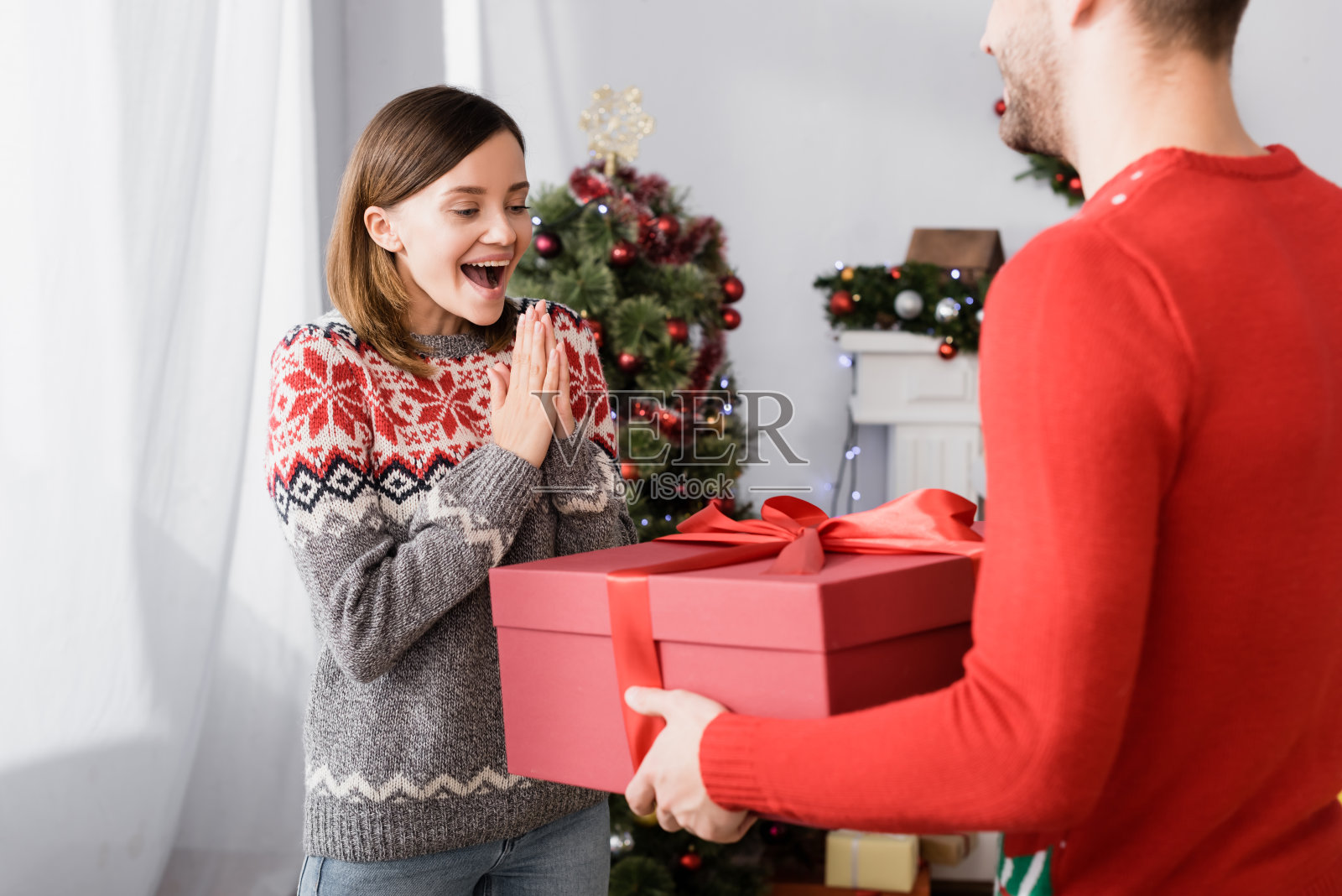 高兴的男人在红色毛衣拿着包裹的礼物近兴奋的妻子和圣诞树在模糊的背景照片摄影图片