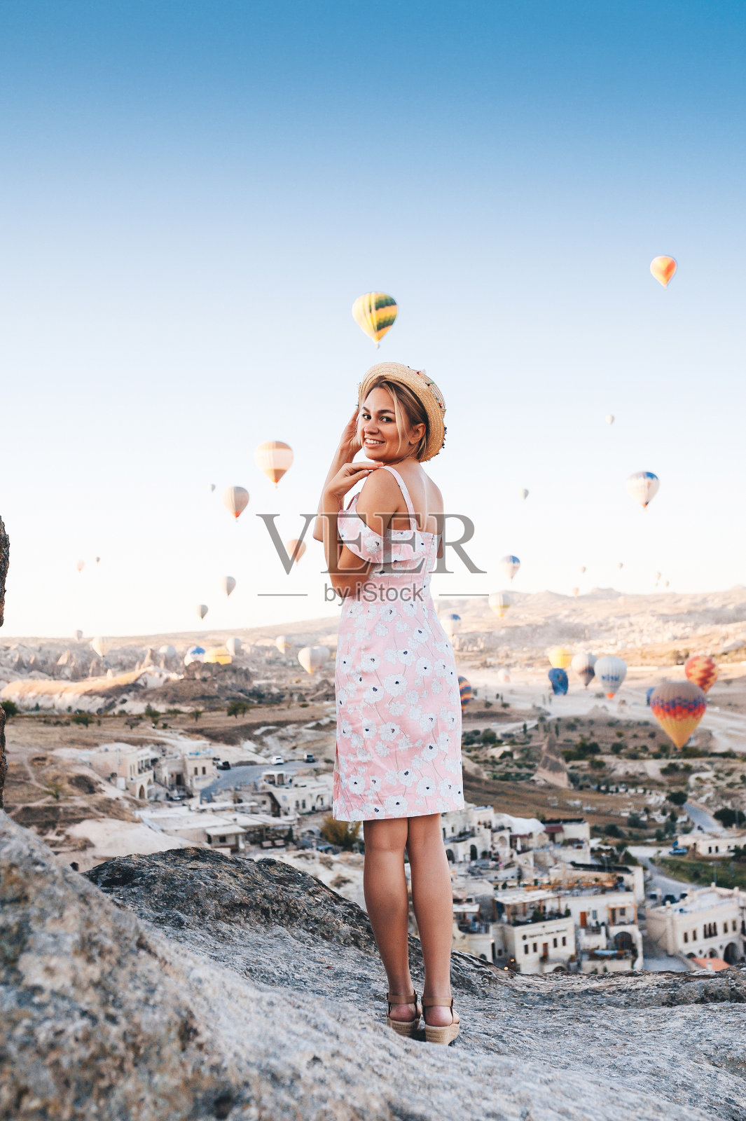 在卡帕多西亚,穿着礼服和帽子的年轻女子在山顶欣赏日出和气球的美妙景色。照片摄影图片