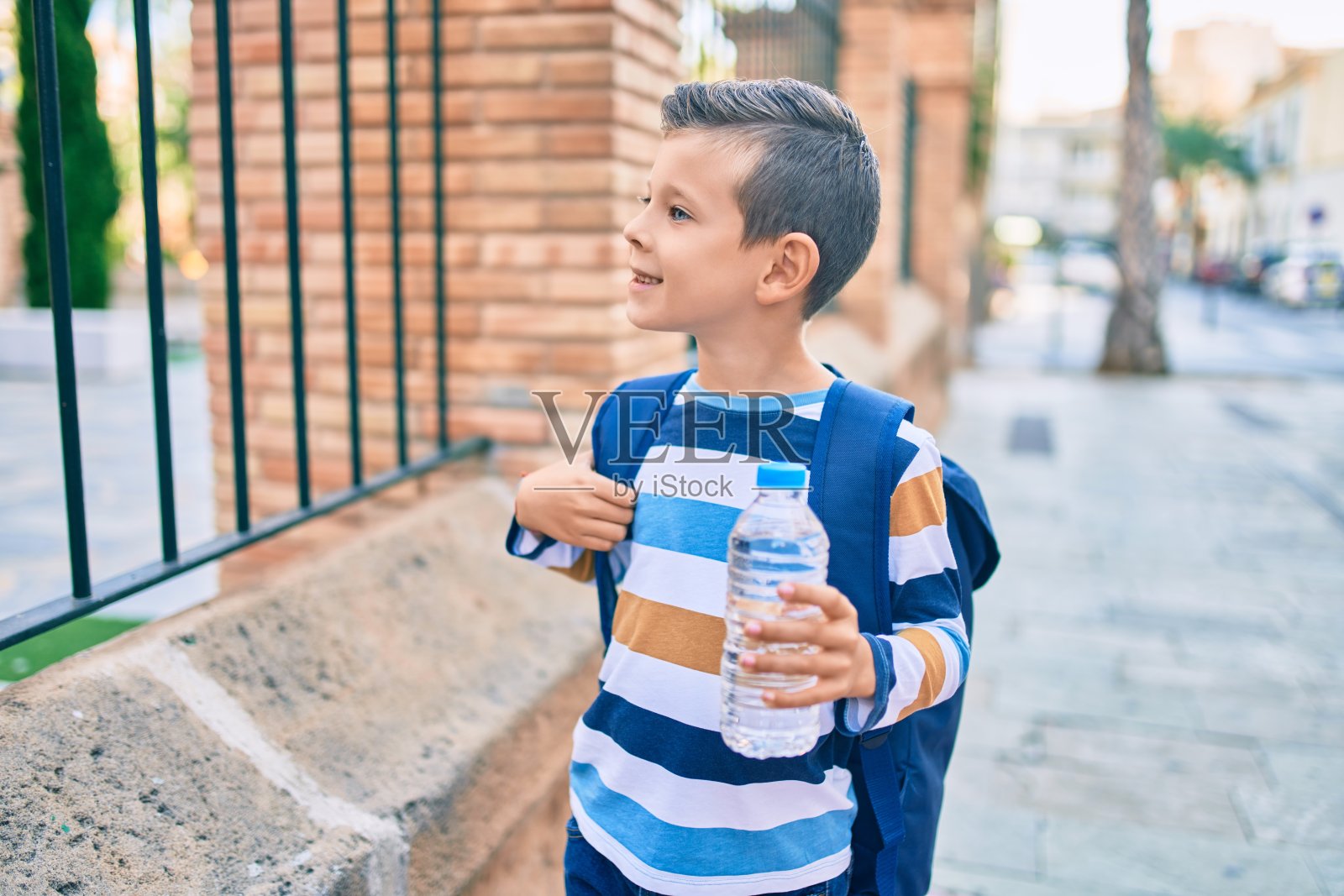 可爱的白人学生男孩开心地拿着一瓶水站在城市。照片摄影图片
