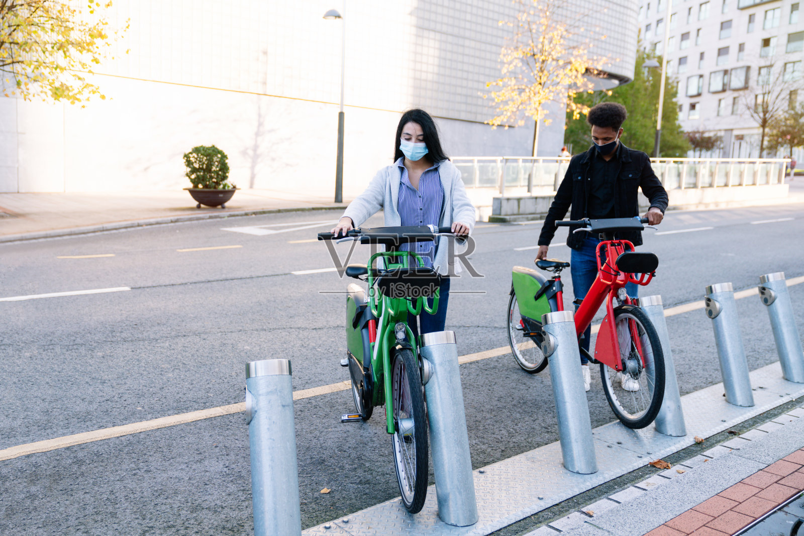 一名摩洛哥青年男子和一名白人女孩戴着口罩在街头自行车停车场租来的电动自行车,以应对2020年新冠肺炎大流行。照片摄影图片