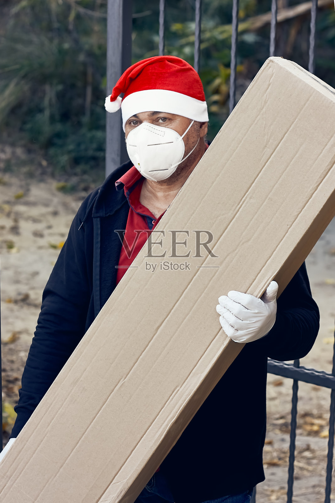 快递员戴着圣诞老人帽、防护面具和医用手套,手里拿着一个大纸箱。速递食品和网上购物的概念。快速交付运输。隔离。照片摄影图片