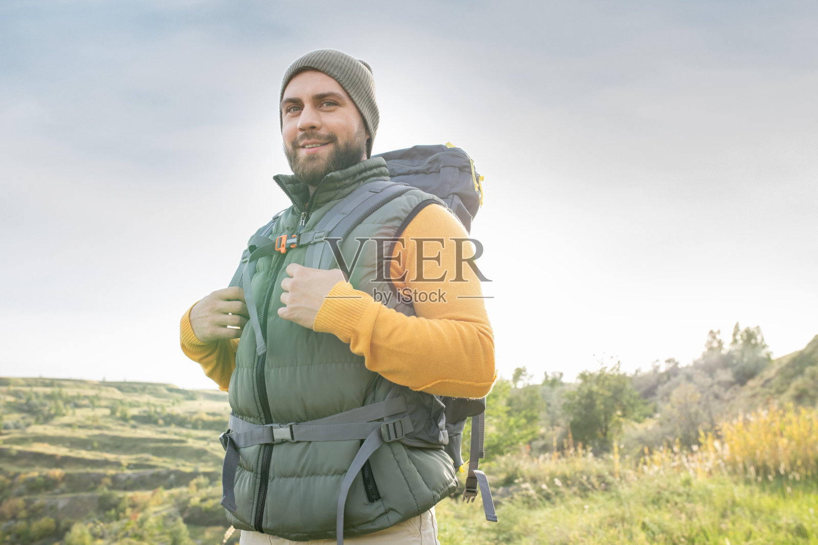 快乐的年轻大胡子背包客在温暖的休闲服站在山照片摄影图片