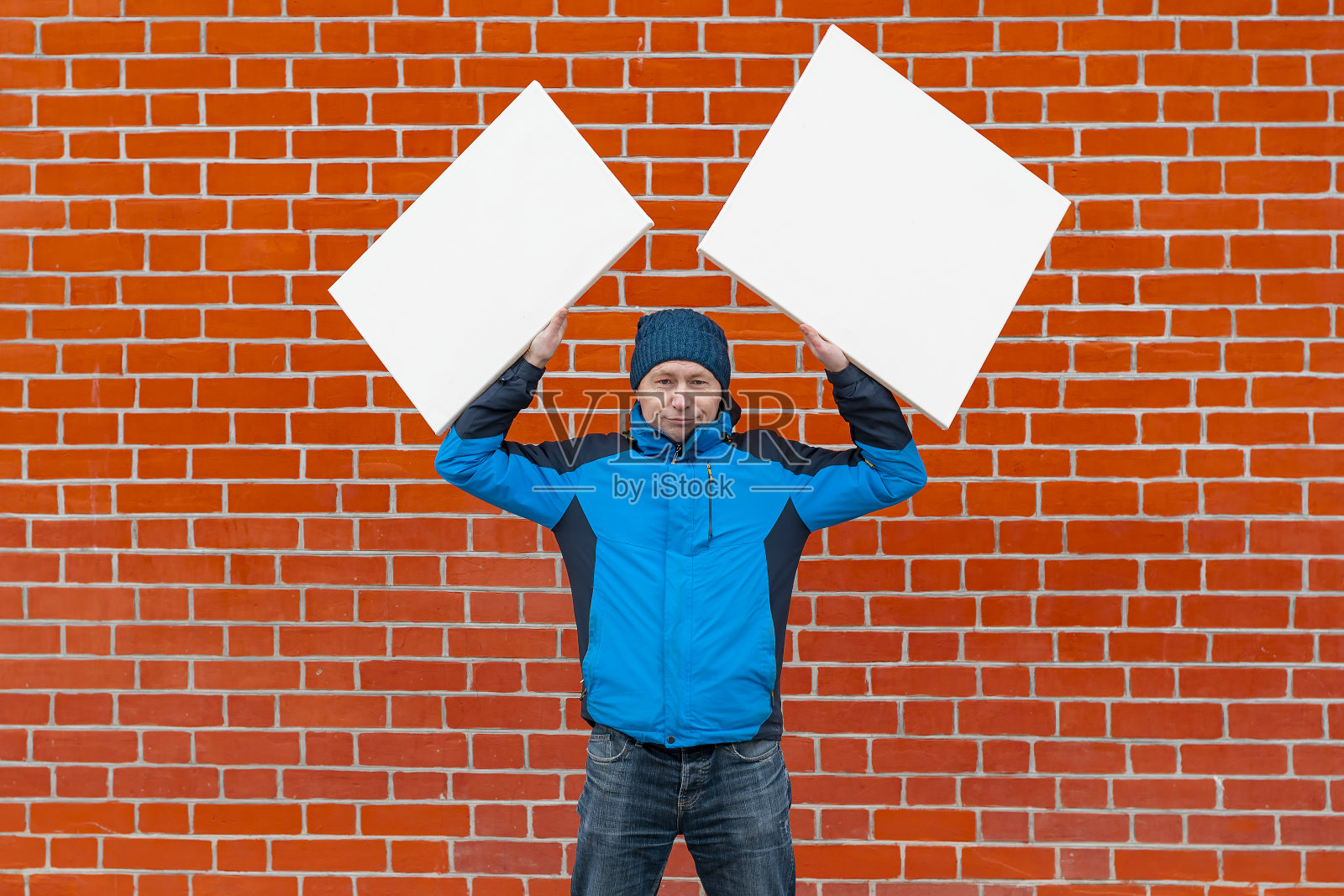 一个年轻人站在红砖墙的背景下,手里拿着两张白色的油画照片摄影图片