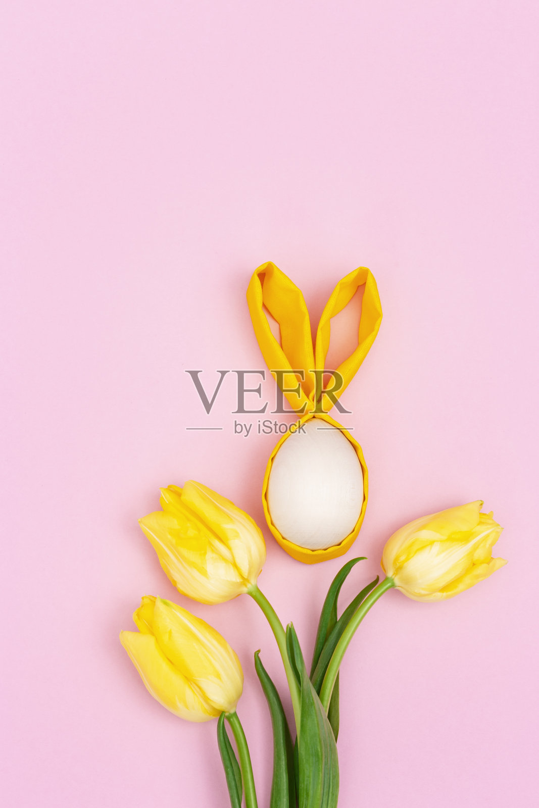 有趣的复活节的概念。一束黄色的花,郁金香和木制的复活节彩蛋与织物耳朵作为有趣的兔子在粉红色的背景。平躺,俯视图。照片摄影图片