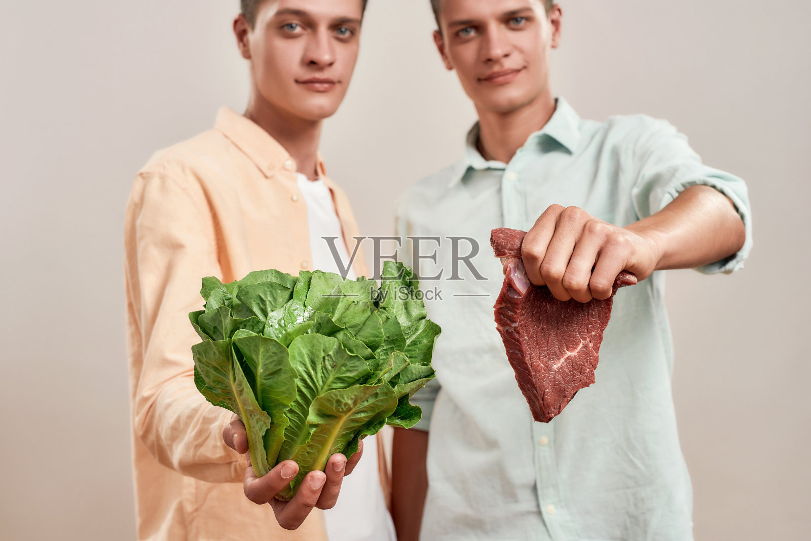 选择健康饮食。两个年轻的白人双胞胎兄弟拿着新鲜的绿色沙拉和一块肉,孤立地站在米色的背景照片摄影图片