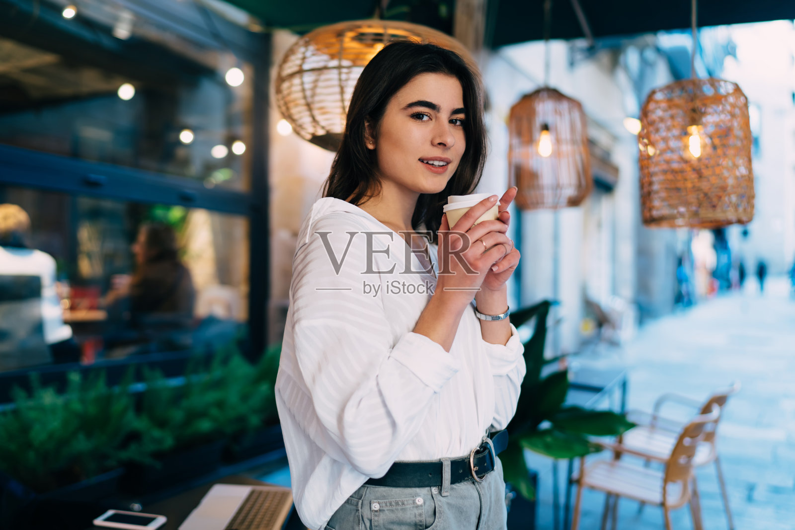 半长肖像,迷人的白人顾客拿着一次性外卖杯,看着相机在街头咖啡休息,穿着随意的时髦女孩与咖啡因饮料摆姿势照片摄影图片