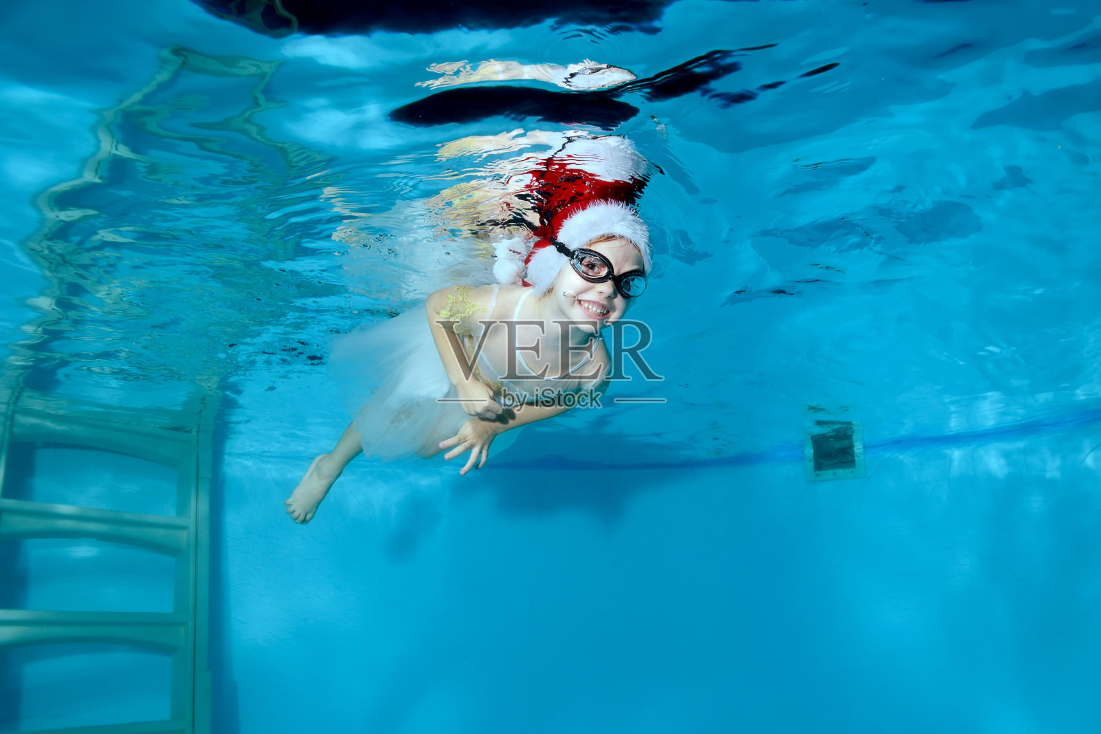 一个戴着圣诞老人帽子和眼镜的笑着的小女孩在儿童游泳池的水下游泳。和一个孩子一起上游泳课。健康的生活方式。水平方向。庆祝的概念。照片摄影图片