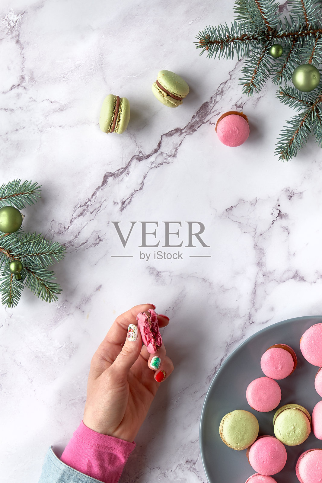 粉色和薄荷绿的杏仁饼干,女人手里拿着吃了一半的通心粉。美味的创意甜点在绿色和红色的大理石桌上与冷杉树枝照片摄影图片