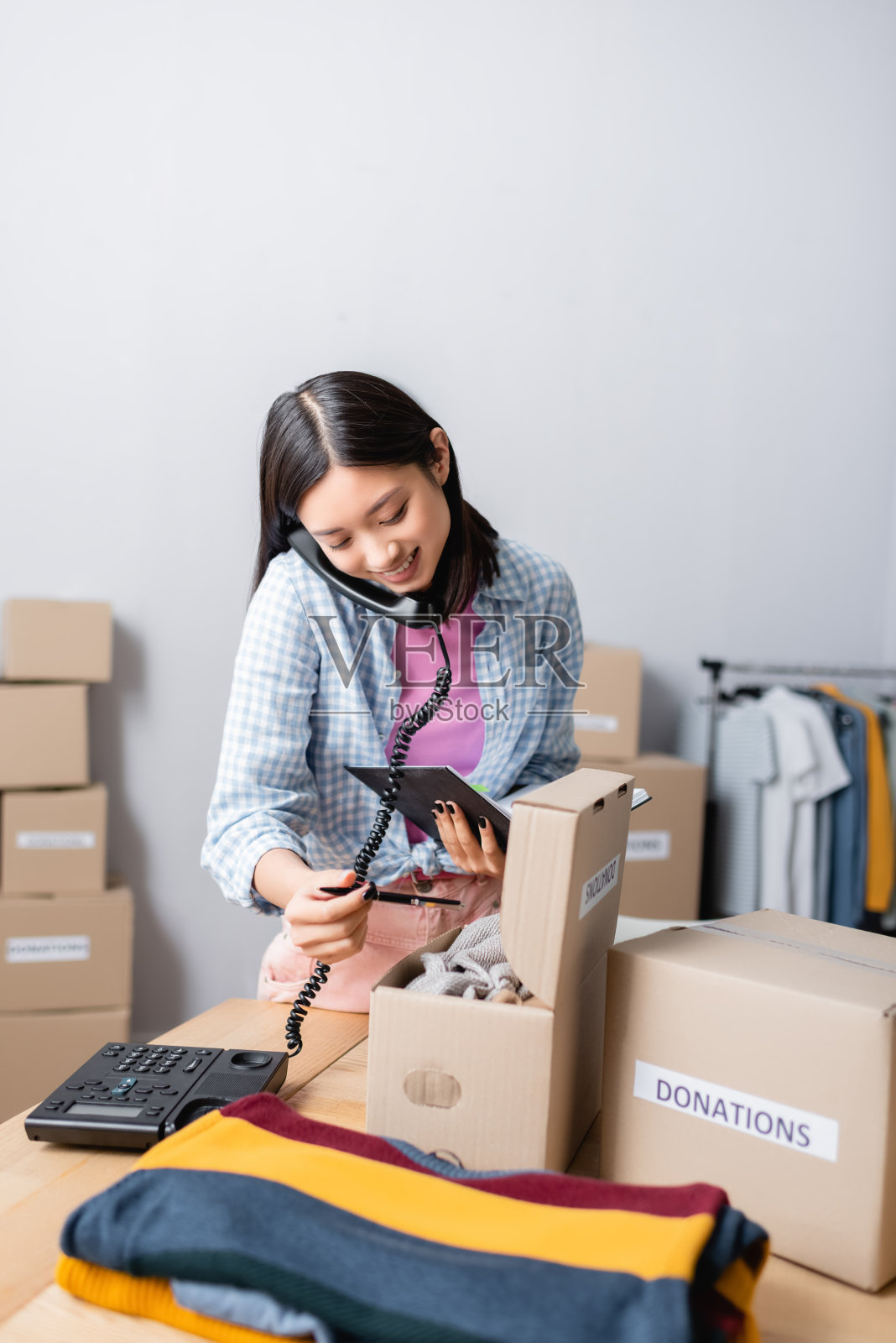 积极的亚洲妇女与笔记本和笔在电话附近与慈善中心的服装盒子照片摄影图片