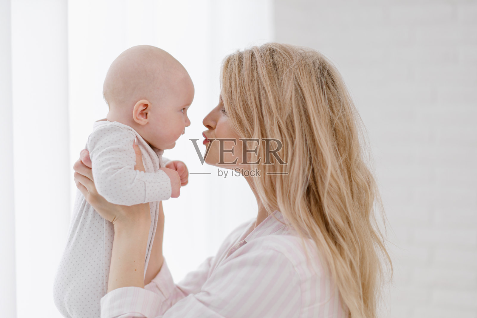 一位年轻的白人母亲穿着家装抱着一个穿着白色连身裤的新生儿。微笑和拥抱孩子。照片摄影图片