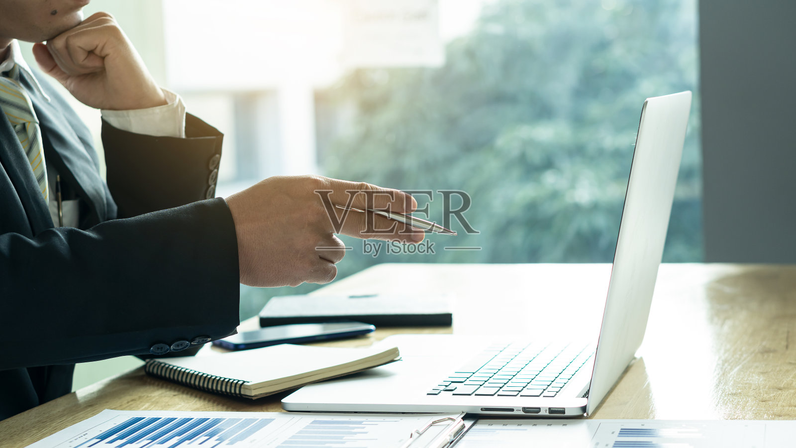 拿一支笔,指着桌子上的商人的笔记本电脑,上面有图表和橙色的灯光。照片摄影图片