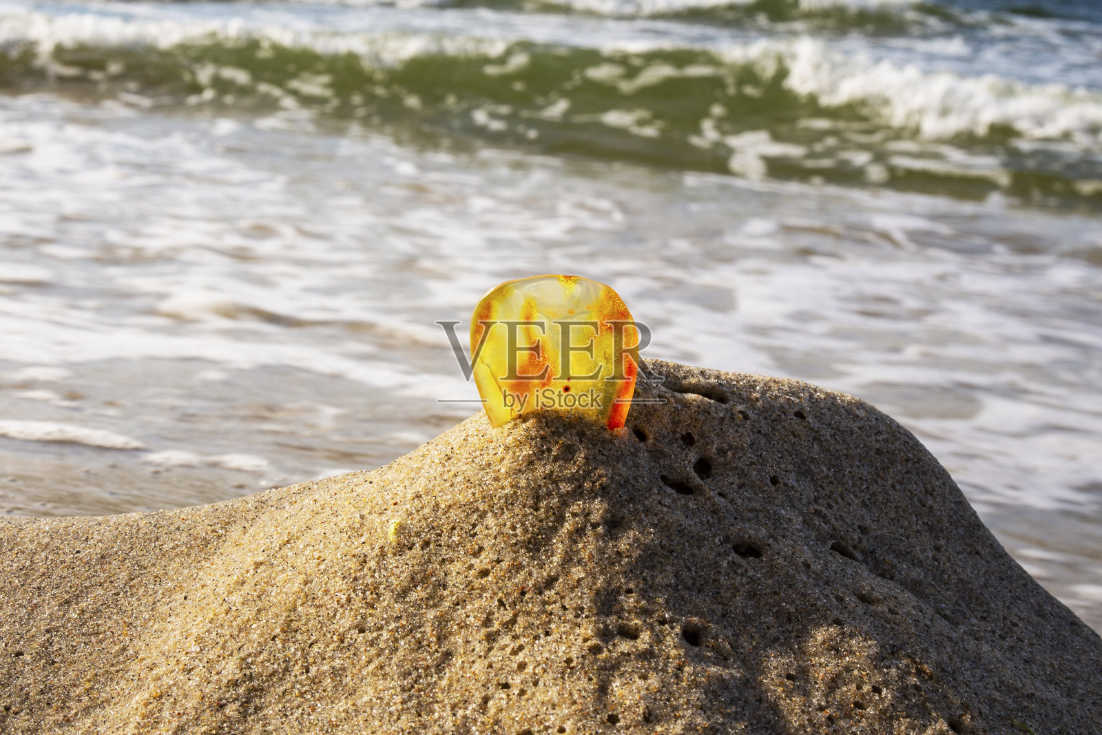 安布尔在沙滩上。一片透明的矿物太阳石在大海的背景上。古老的树脂,在沙滩上加工过的琥珀,背景中模糊了海浪的背景。波罗的海照片摄影图片