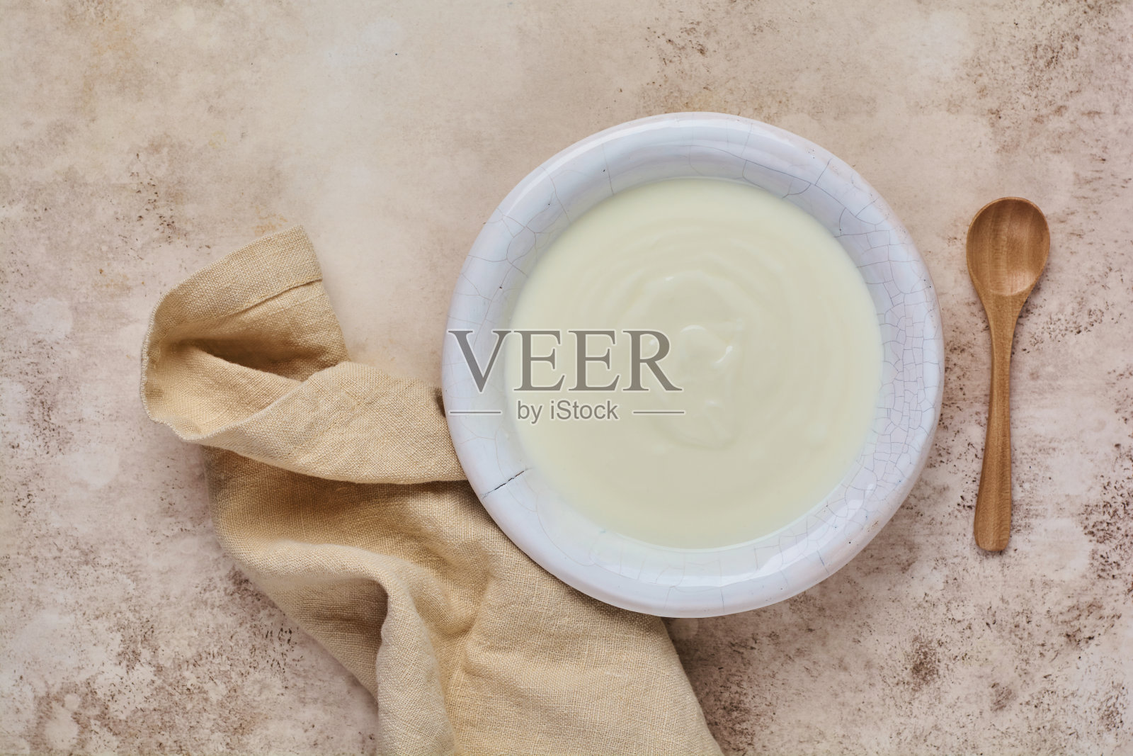白碗里的希腊酸奶,亚麻餐巾和木勺放在米色的旧桌子上。前视图。照片摄影图片