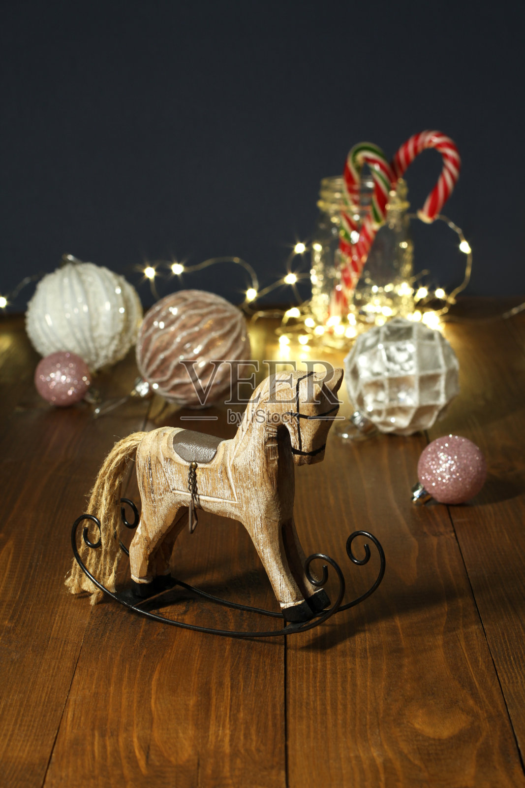 圣诞装饰品——木制玩具摇木马,圣诞树球,玻璃罐里的新年糖果,在蓝色墙壁边的木桌上,一个发光的花环缠绕着它照片摄影图片