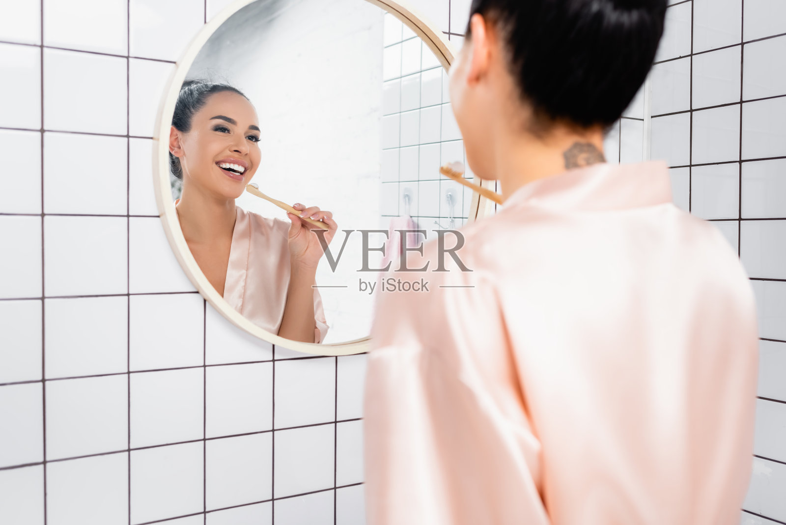 穿着缎子浴袍的微笑女人看着镜子,手里拿着牙刷在浴室模糊的前景照片摄影图片