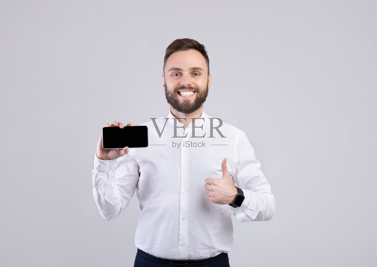 酷酷的蓄着胡子的家伙展示着空屏幕的智能手机,并在工作室的灰色背景上竖起大拇指照片摄影图片