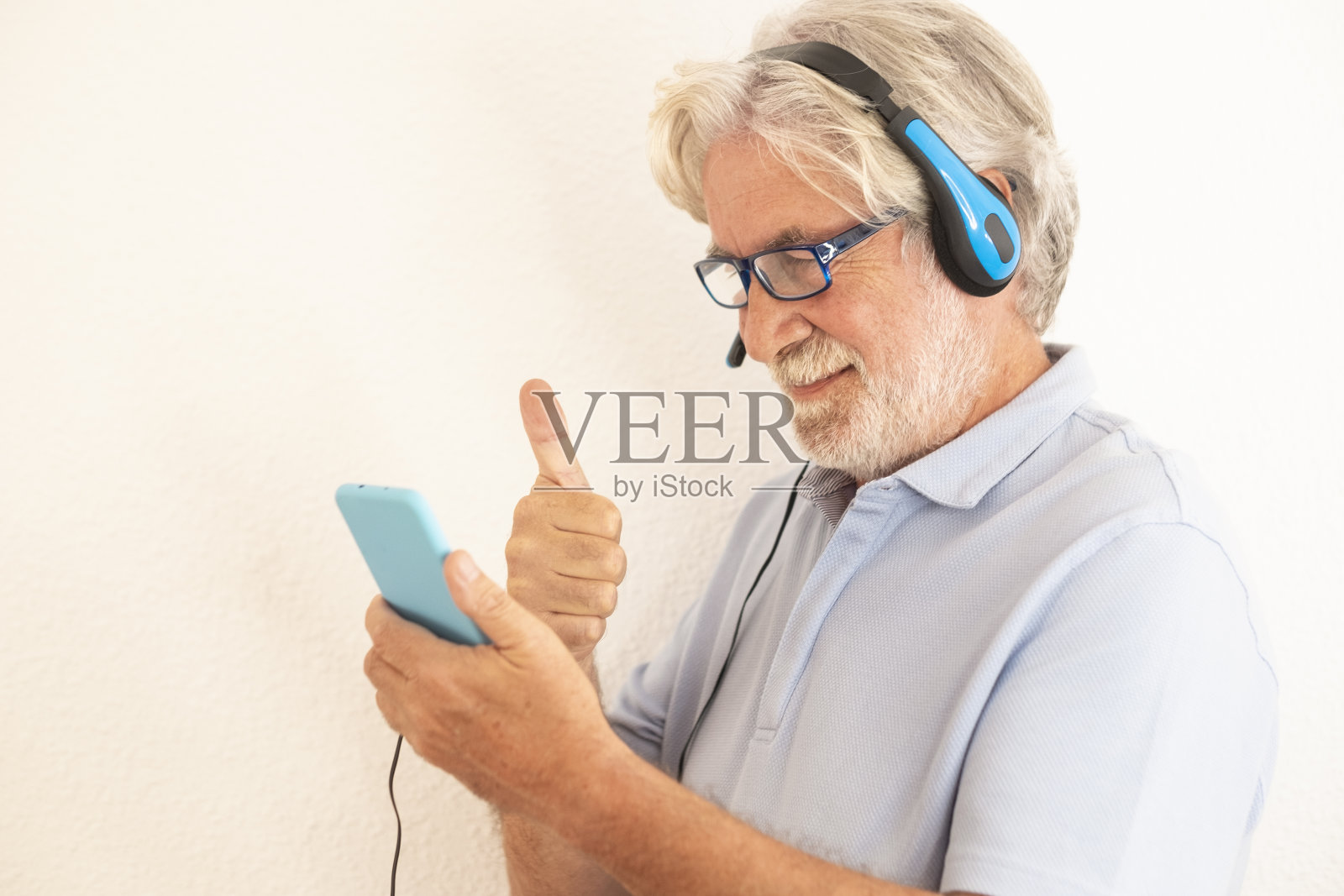白发老人戴着耳机站在白色背景下看他的智能手机与家人或朋友视频通话。老年人享受新技术照片摄影图片