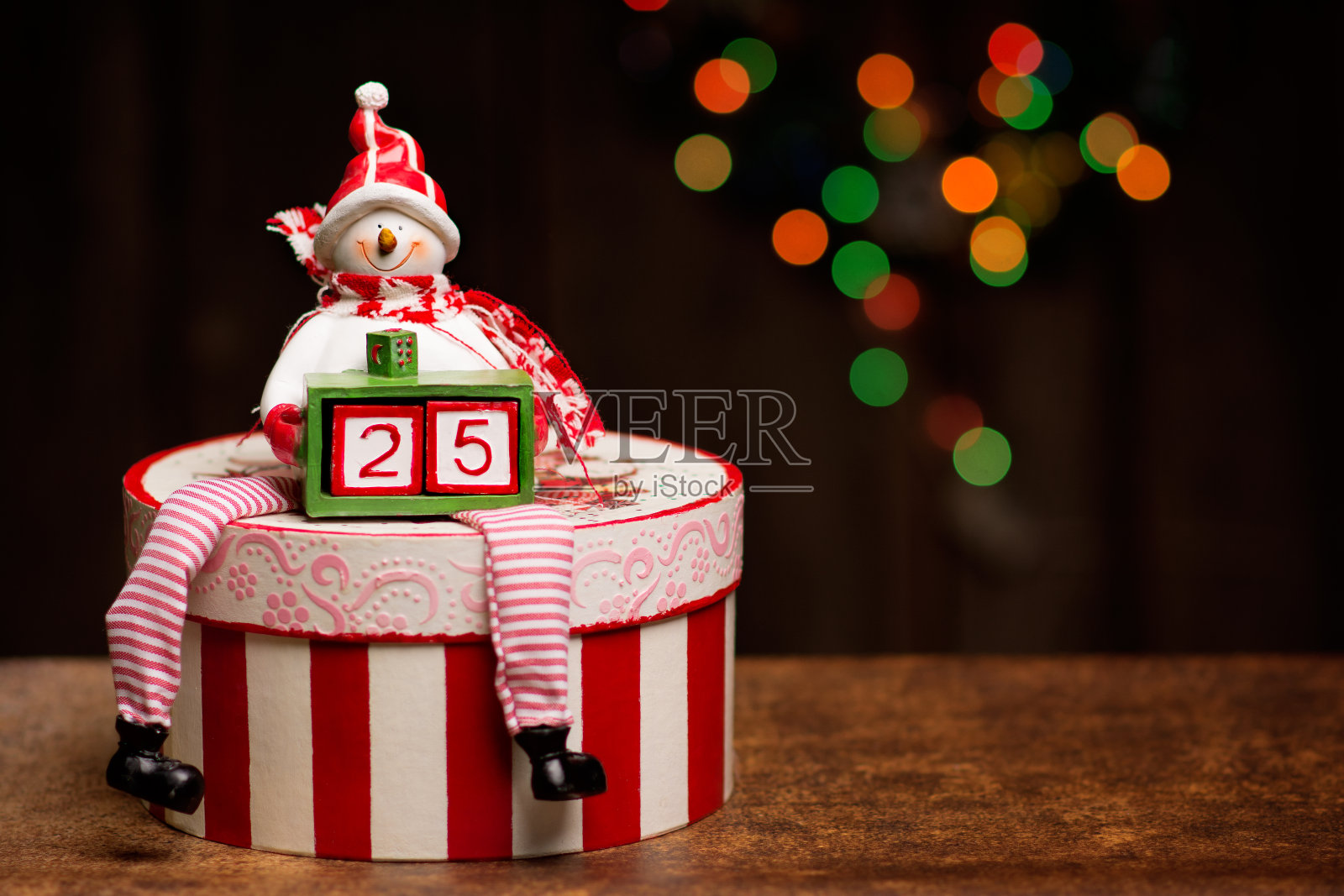 一个雪人正坐在一个装饰精美的剪纸盒子上,手里拿着一个小盒子,盒子里有两个立方体组成数字25。一个美丽的散景与圣诞灯是在背景。照片摄影图片