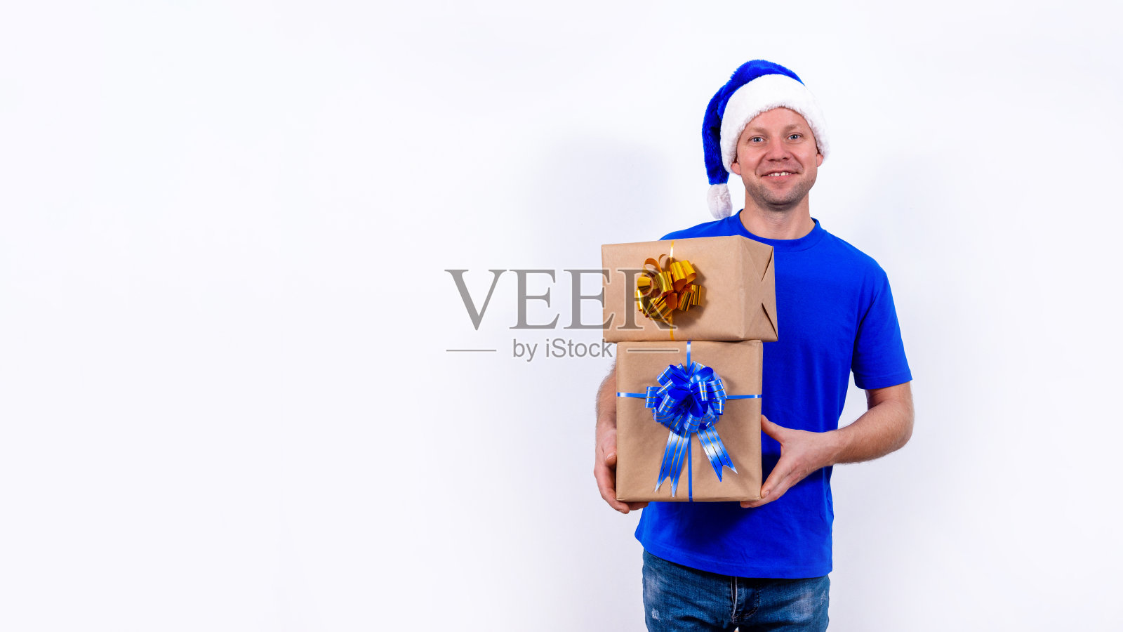 横幅。一个快乐的信使穿着蓝色的制服和圣诞老人的帽子拿着许多礼物盒。新年快乐,圣诞快乐。那人微笑。安全的非接触远程递送节日礼物。照片摄影图片