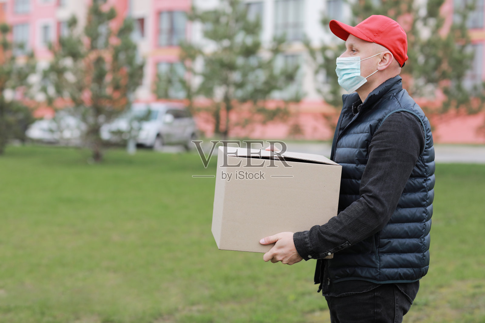 送货人戴着红帽子,戴着医用口罩,手持空纸板箱在户外。服务的冠状病毒。在线购物。模拟。照片摄影图片