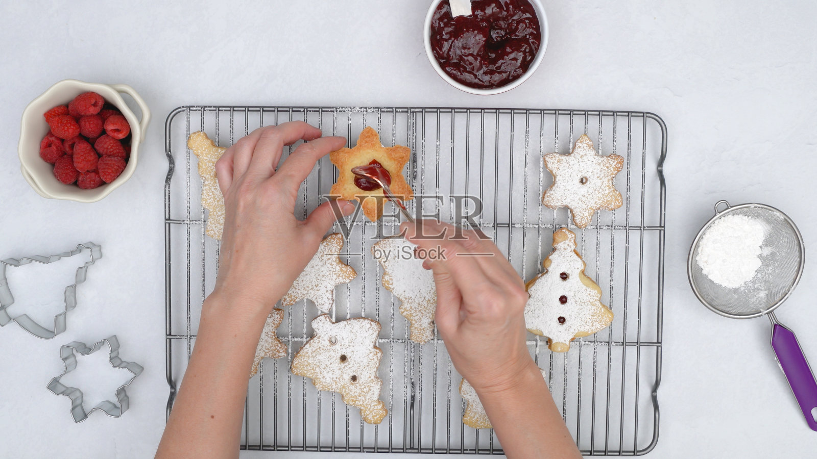 圣诞酥饼配覆盆子果酱食谱,封闭烘焙过程。烤饼干放在冷却架上,女人用手在饼干上抹上一层覆盆子果酱照片摄影图片