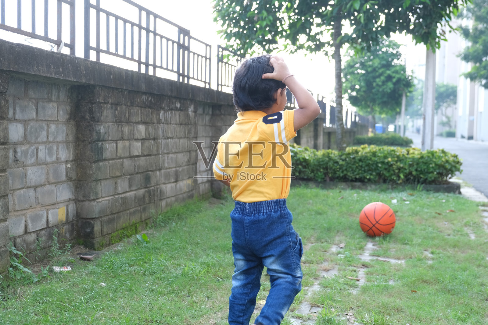 印度可爱的小愉快的深褐色的泰米尔婴儿男孩在充满活力的黄色休闲服玩自己在绿色自然背景的一个橙色球。照片摄影图片