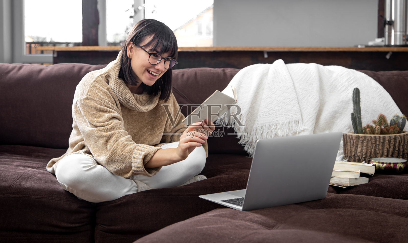 一个戴着眼镜,兴高采烈的年轻女人,手里拿着一本书,坐在家里沙发上的笔记本电脑后面。照片摄影图片