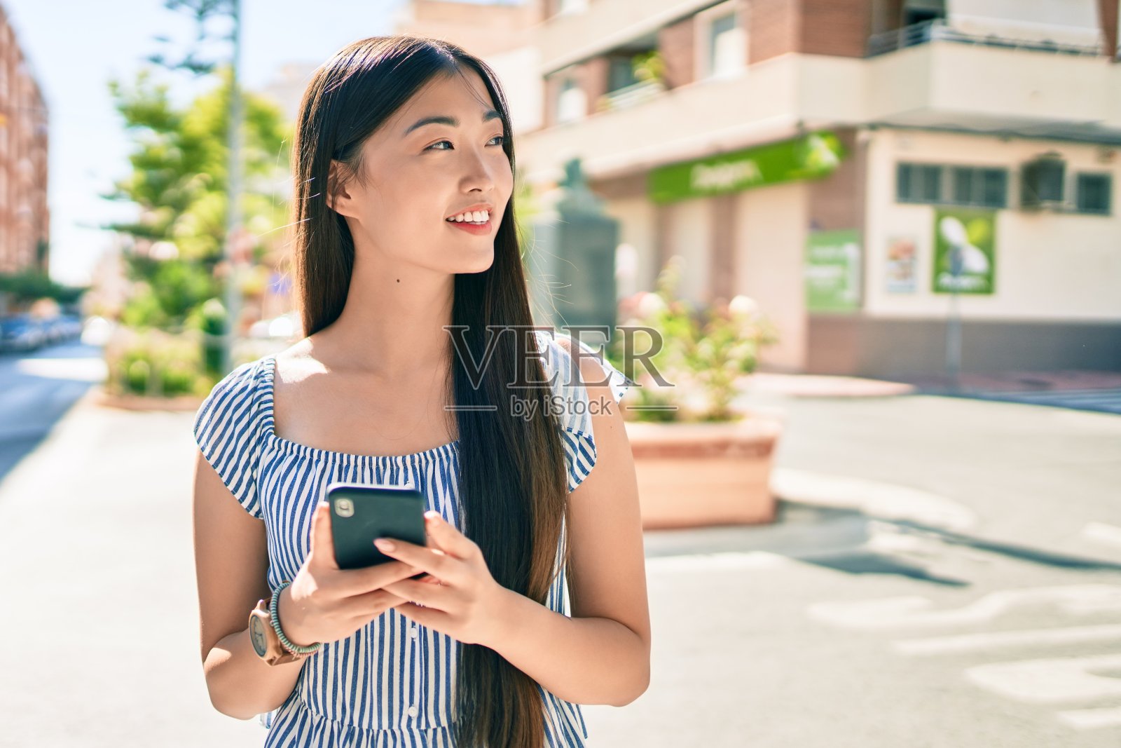 年轻的中国女子在城市的街道上用智能手机微笑。照片摄影图片