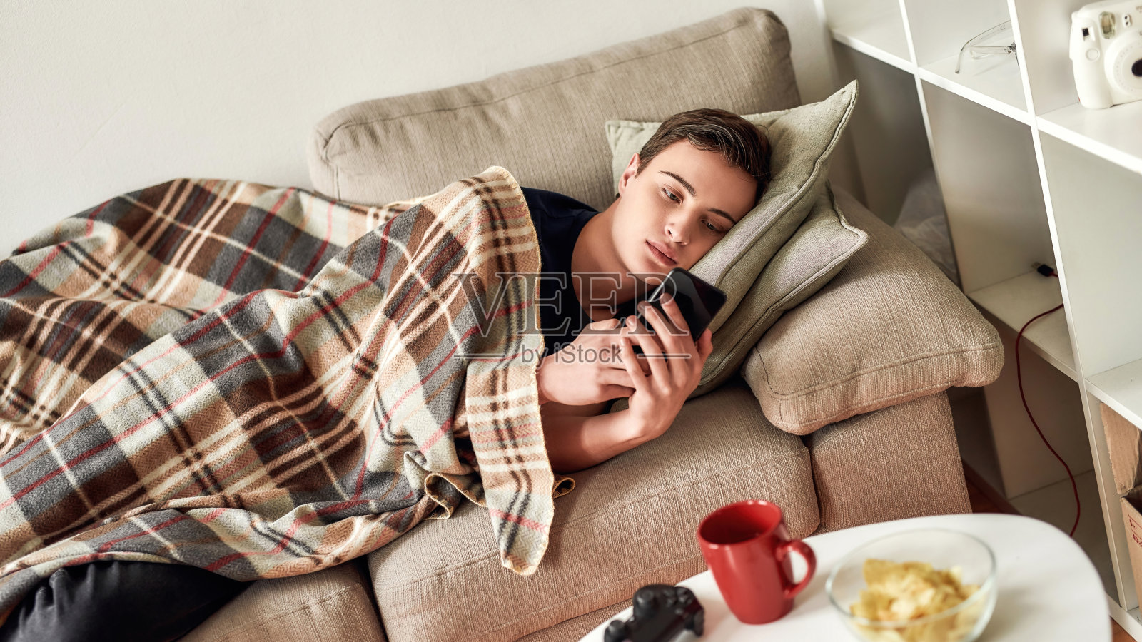 一个十几岁的男孩躺在家里的沙发上休息的时候用手机。对智能手机上瘾、冷漠和沮丧的概念照片摄影图片