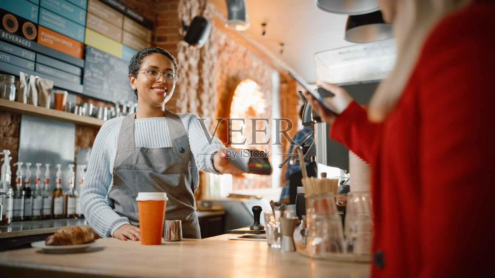 快乐的多种族不同的女人在智能手机上使用NFC技术给客户一个支付终端。顾客使用手机为咖啡店的咖啡师支付外卖拿铁和羊角面包。照片摄影图片