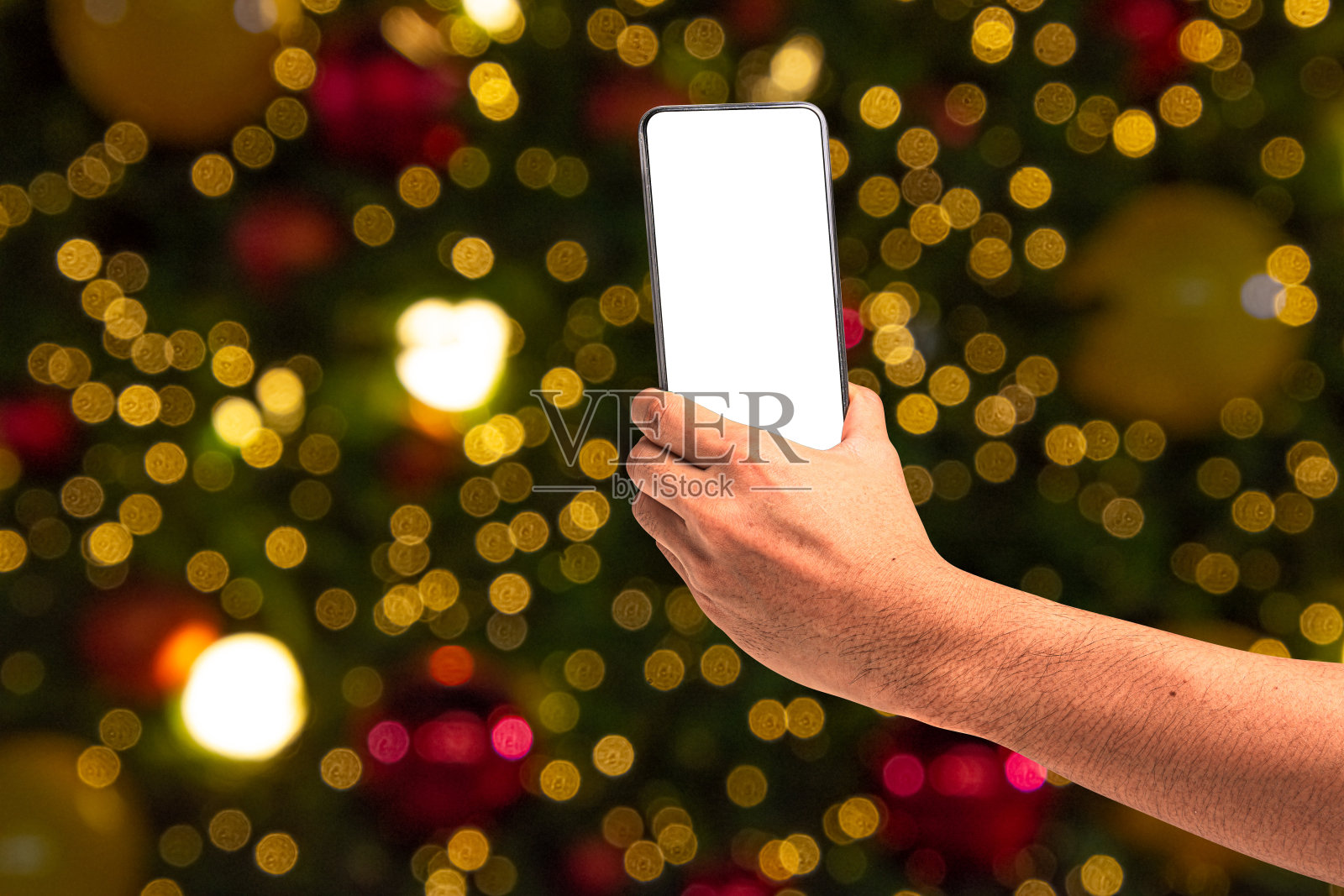 近距离手持手机孤立在白色背景与照明模糊的散景圣诞树装饰与悬挂灯球。照片摄影图片