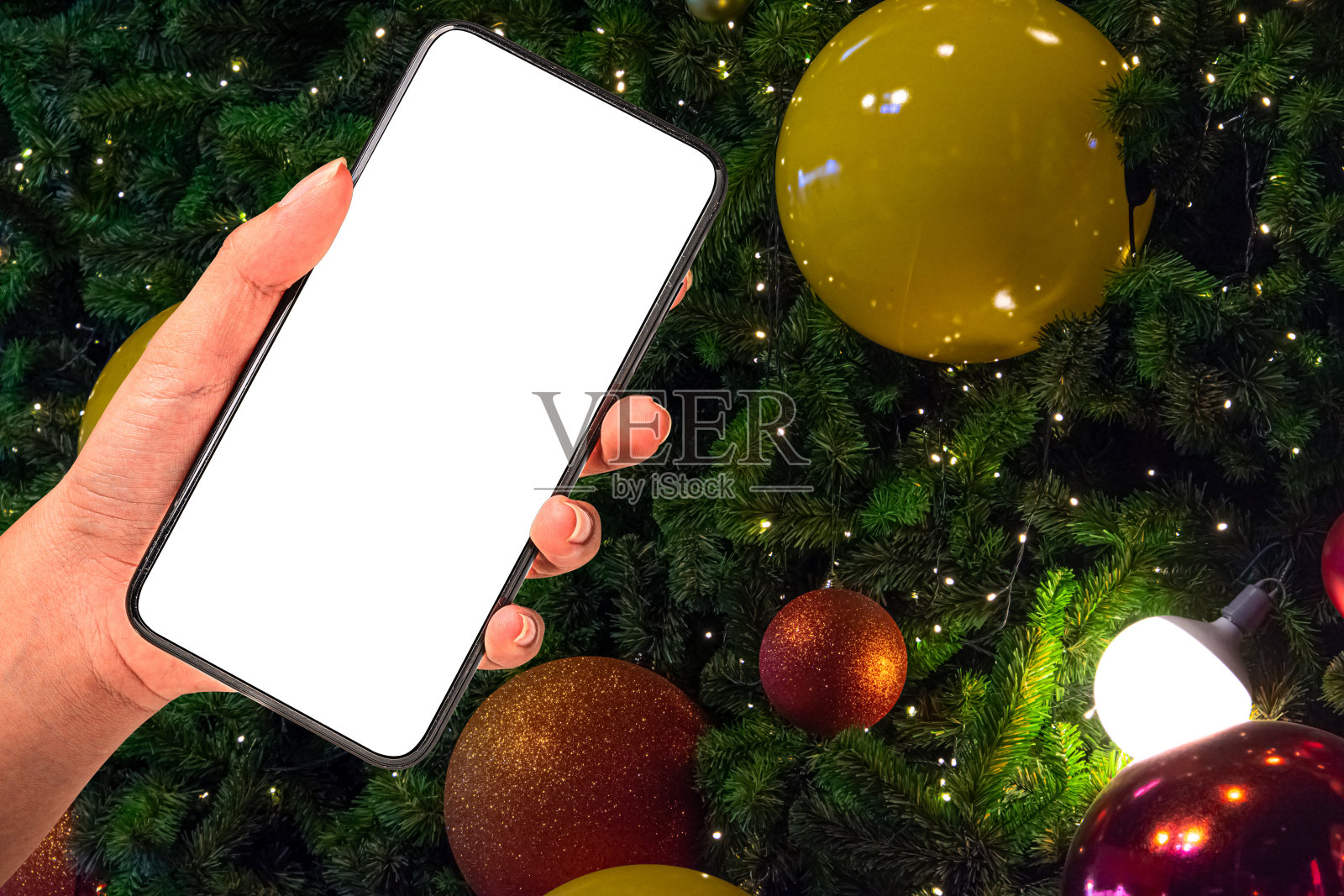 近距离手持手机孤立在白色的背景与灯光装饰圣诞树挂着黄色和红色的球。照片摄影图片