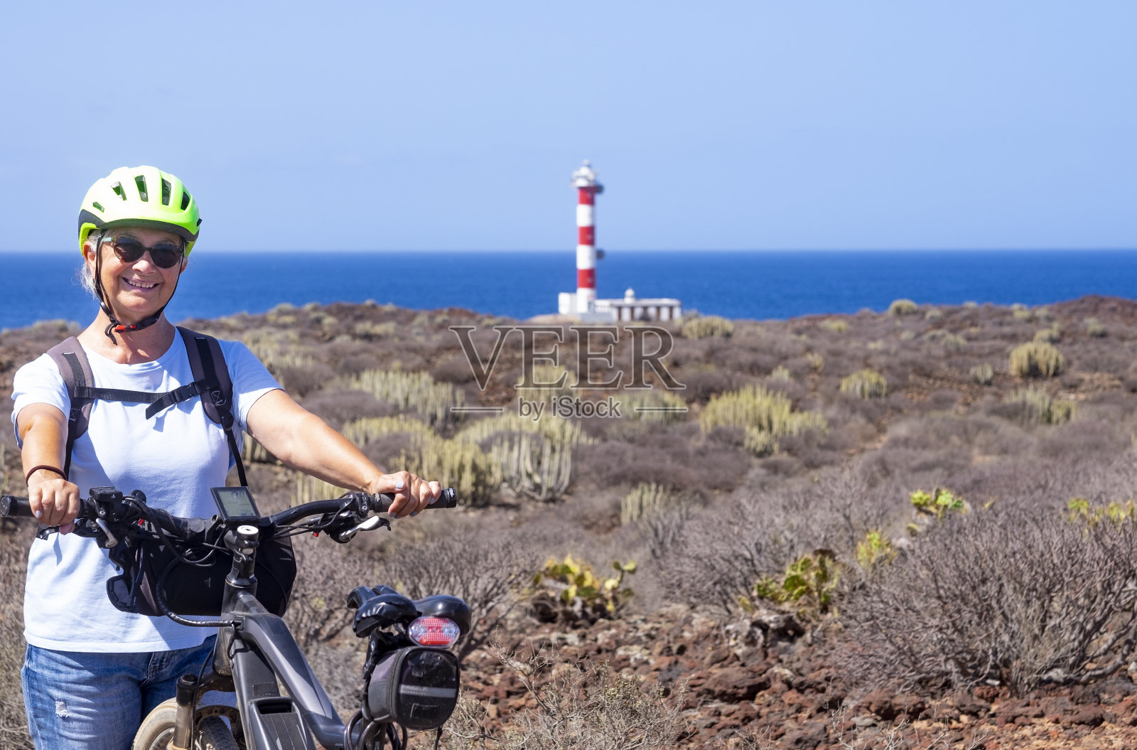 快乐的妇女与黄色头盔享受自由和远足与自行车靠近灯塔-干旱的景观和地平线上的水在一个阳光明媚的一天在特内里费,加那利群岛照片摄影图片