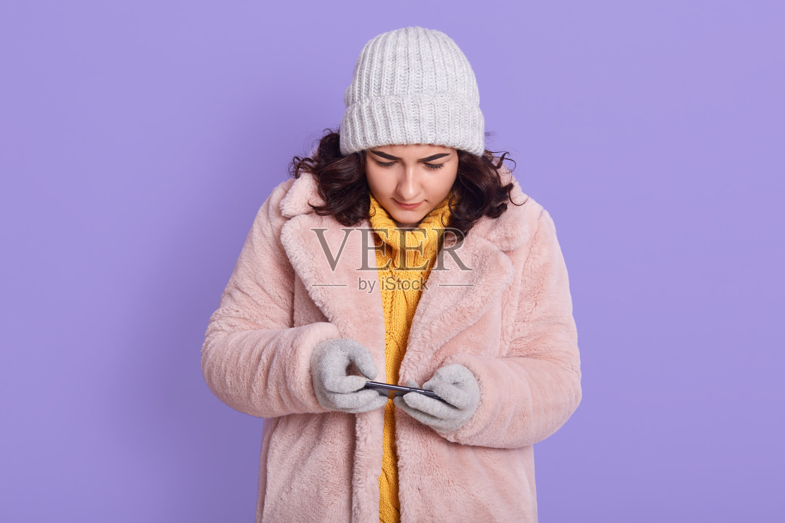 年轻美丽专注的女人在淡紫色的墙壁背景下摆出孤立的姿势,玩手机游戏,看展示,穿着人造皮草大衣,帽子和手套。照片摄影图片