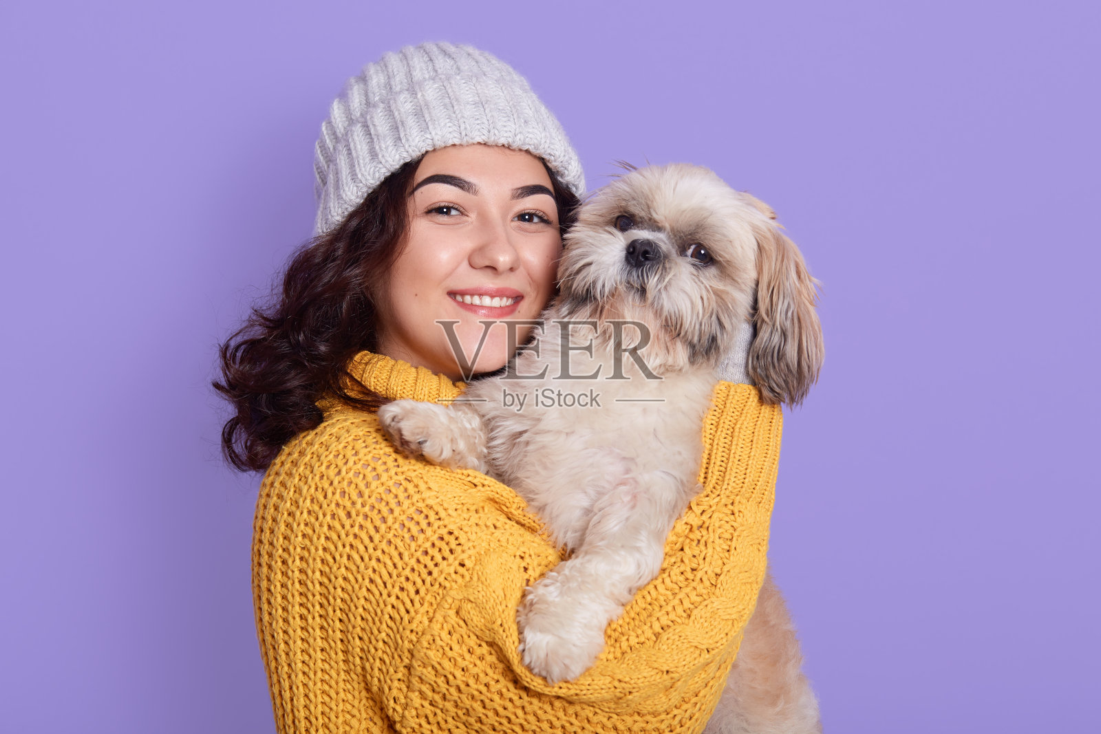 迷人的女性,有着可爱的外表,在和北京小狗拍照时表达积极的情绪,温柔地拥抱近脸,直视镜头微笑,穿着针织毛衣和帽子。照片摄影图片