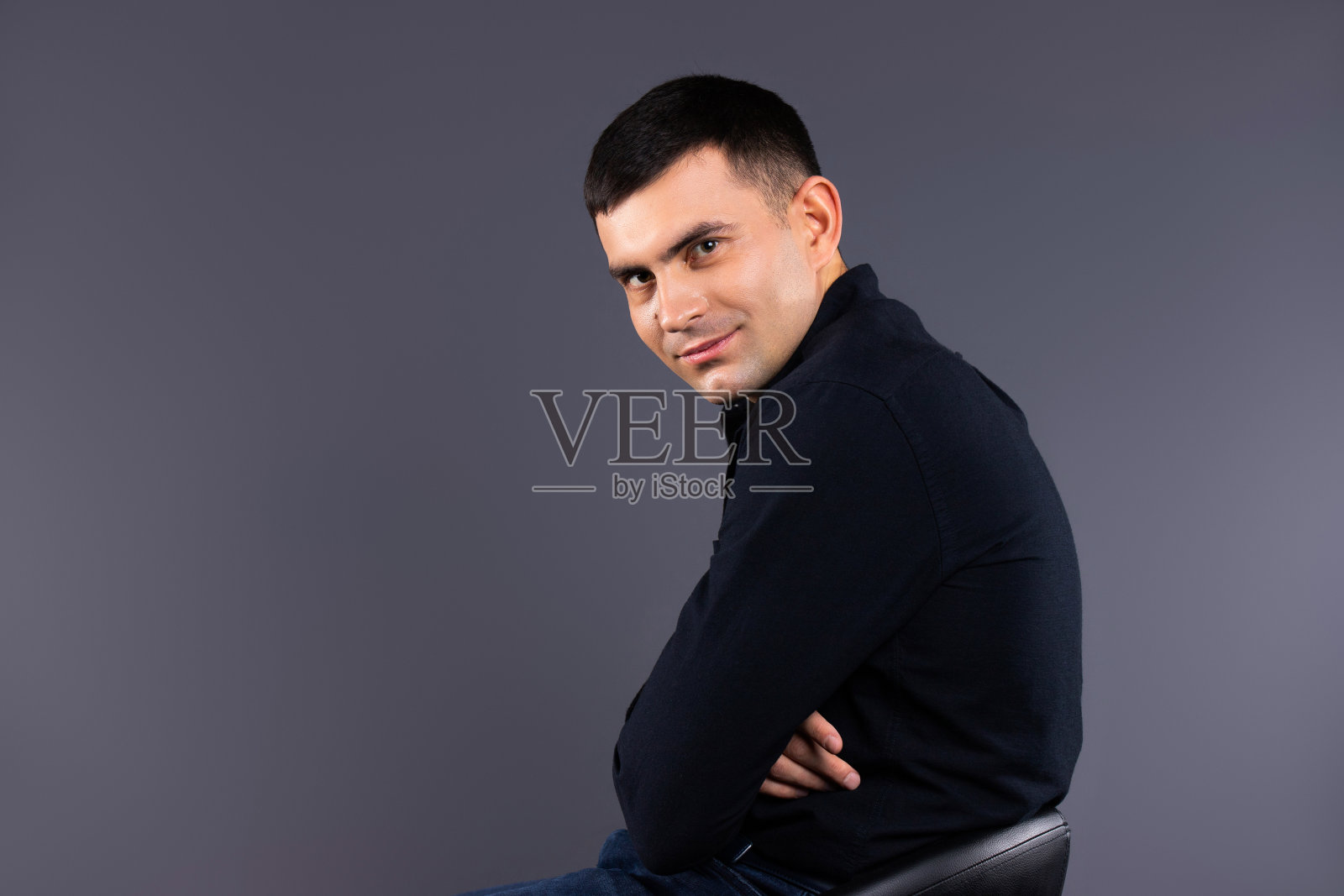 一名穿着黑色衬衫、坐在真皮椅子上、灰色背景的时尚男子看着摄像机。档案照片。照片摄影图片