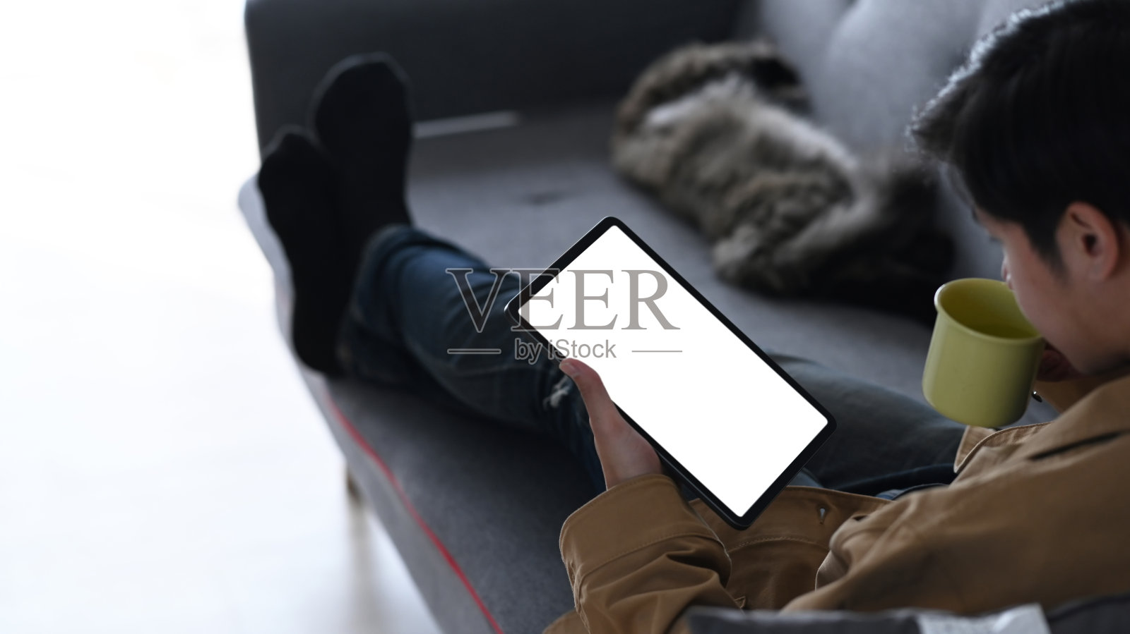 一个年轻人用着平板电脑,喝着咖啡,和他的猫在沙发上休息。照片摄影图片