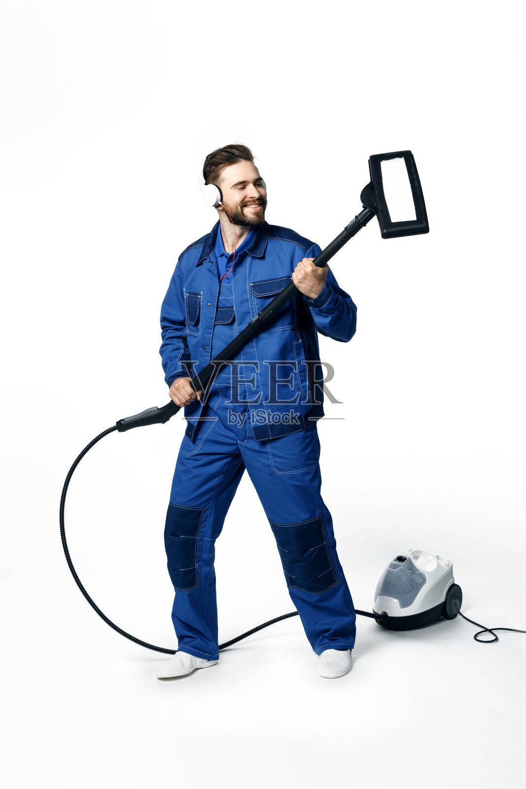 年轻英俊的男子留着胡子,穿着蓝色工作制服打扫房间,戴着耳机听音乐,微笑着在白色背景下吸尘照片摄影图片