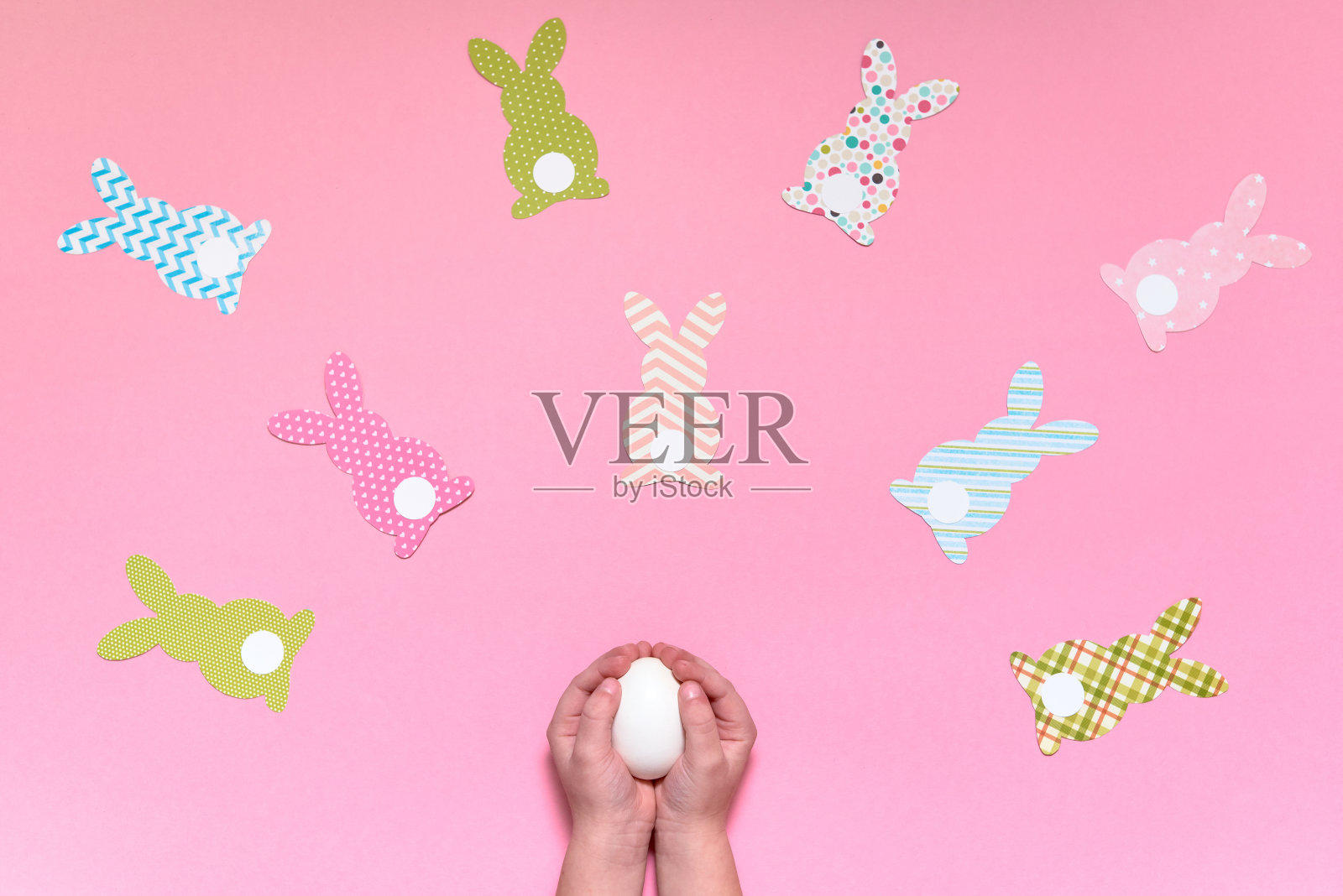 孩子手里拿着白色的蛋在粉红色的背景与复活节兔子装饰,复制空间。DIY节日手工艺品兔子。平铺,俯视图。复活节贺卡。复活节快乐概念照片摄影图片