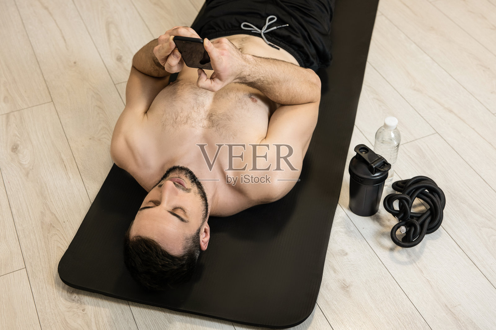 健身者在努力健身后休息,并在电话上与朋友发短信。运动器材。休息时间。懒人躺在黑色瑜伽垫上,用智能手机发短信。照片摄影图片